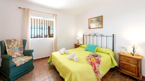 A bedroom with a bed, a chair, a nightstand, a window with bars, and a painting on the wall. - Villa Sanchez Sol by At Home Costa del Sol (Torrox)