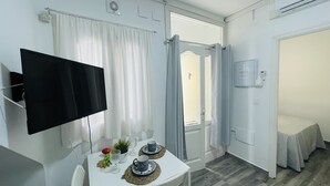 Ein kleiner, sauberer Raum mit einem Flachbildfernseher, einem Esstisch mit Blumenvase und einer Tür zu einem weiteren Zimmer.
