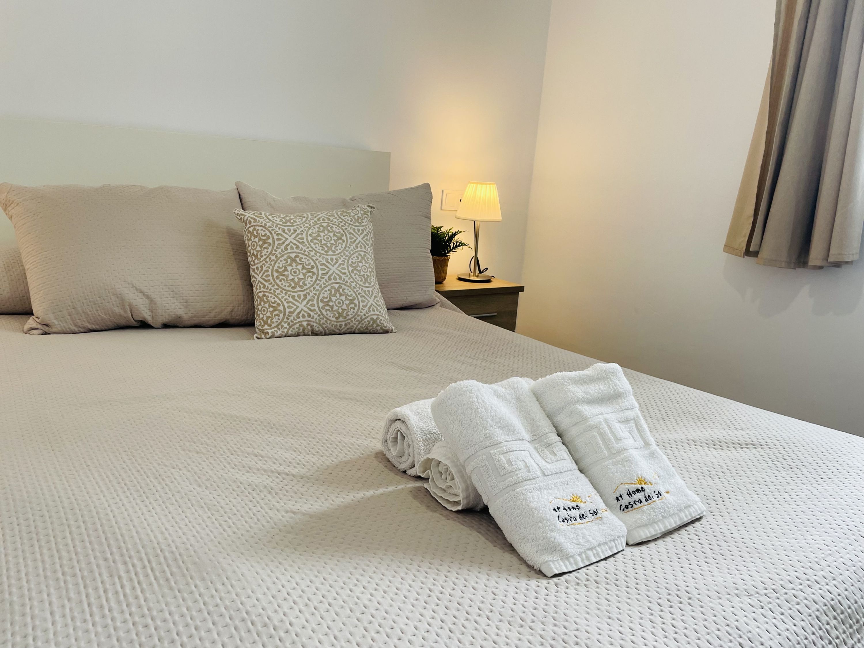A neatly made bed with a patterned pillow, a bedside lamp, and a folded towel with text on it.