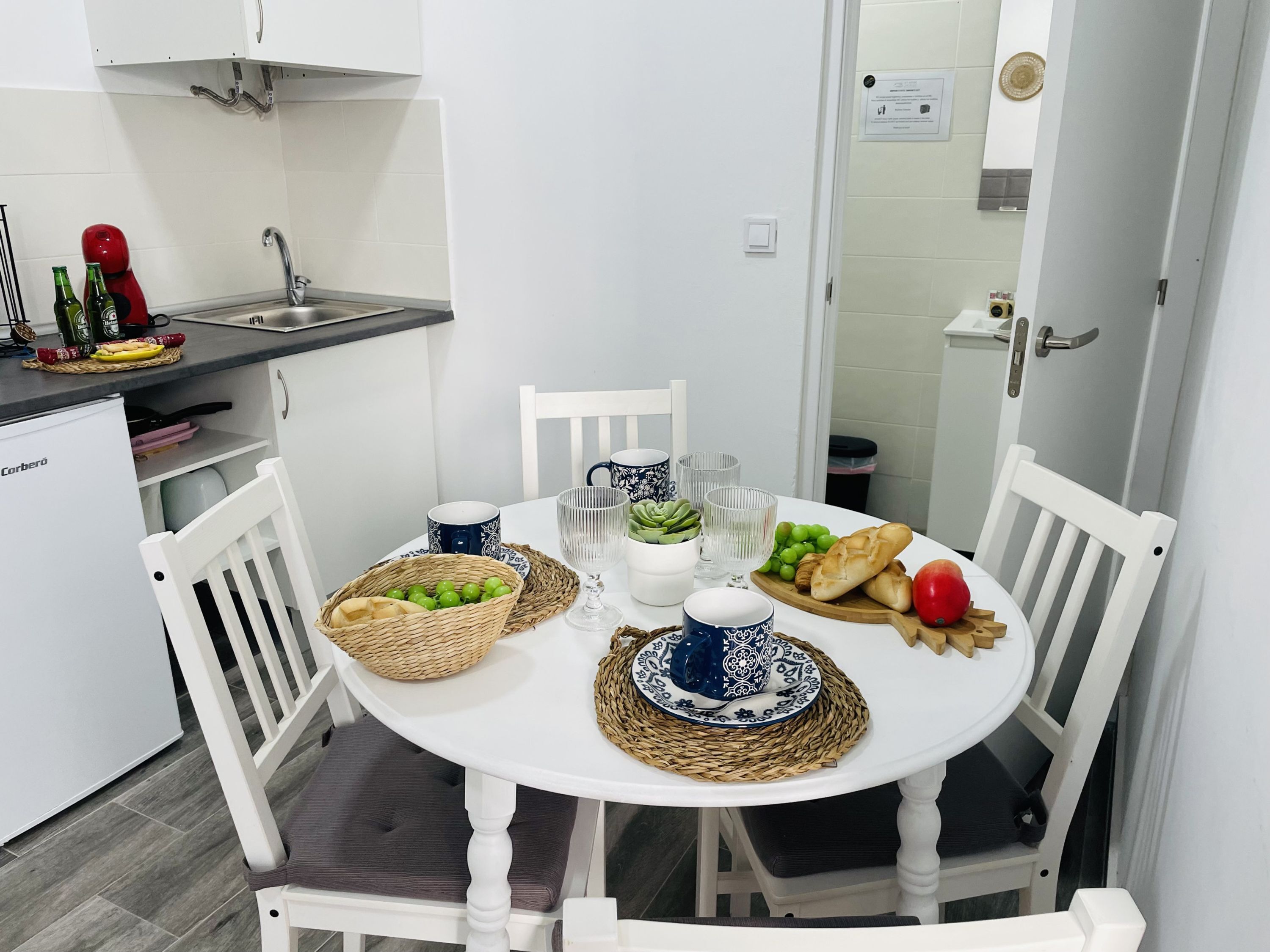 Een praktische keuken met witte kastjes, een spoelbak en een kleine koelkast. Een eettafel met witte stoelen is gedekt met borden, kommen en een mand vol druiven.