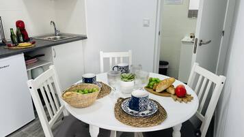 Een praktische keuken met witte kastjes, een spoelbak en een kleine koelkast. Een eettafel met witte stoelen is gedekt met borden, kommen en een mand vol druiven.