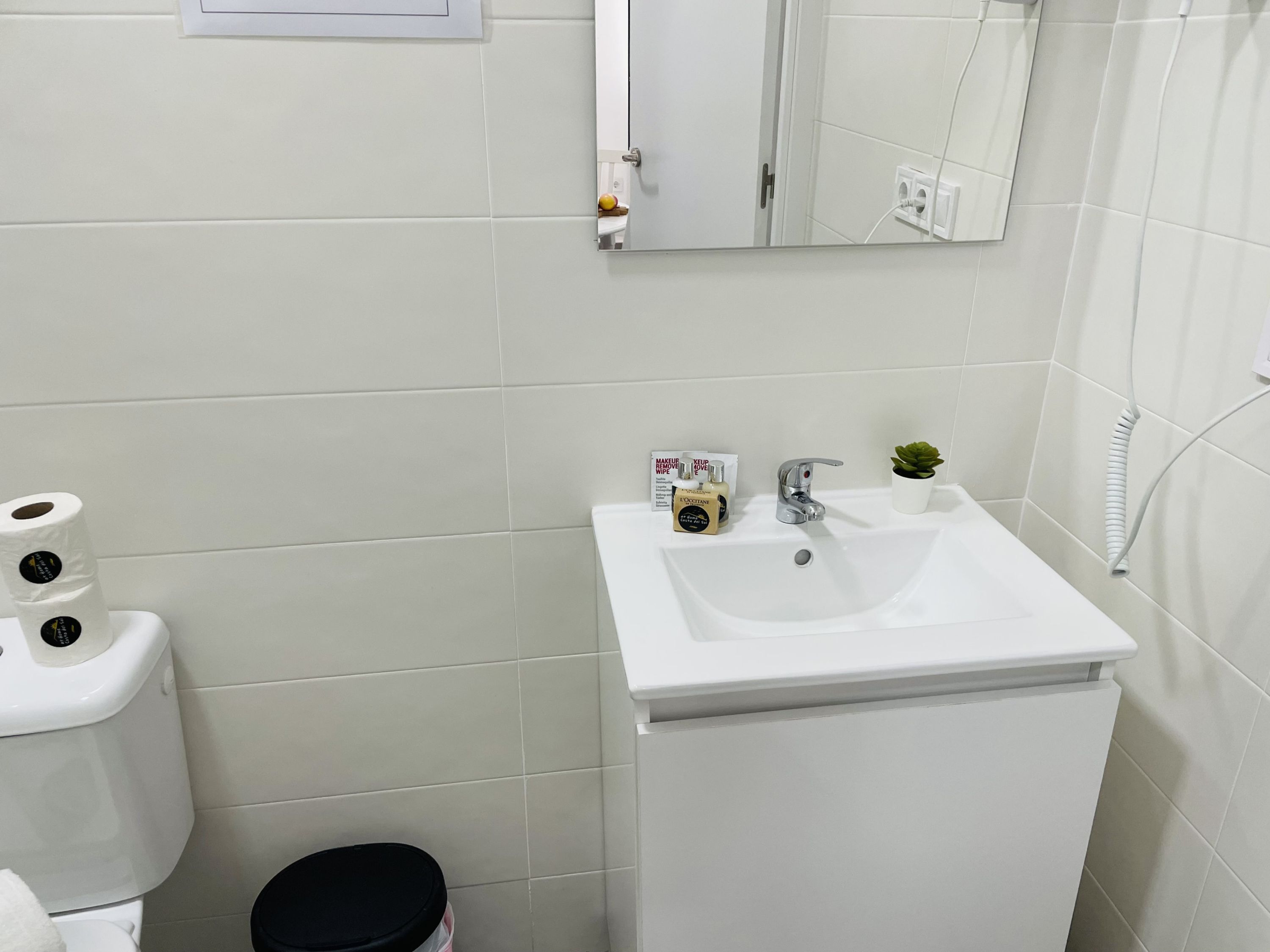 Ein Badezimmer mit weißem Waschbecken, einem Spiegel, einer Toilette und einem Mülleimer.