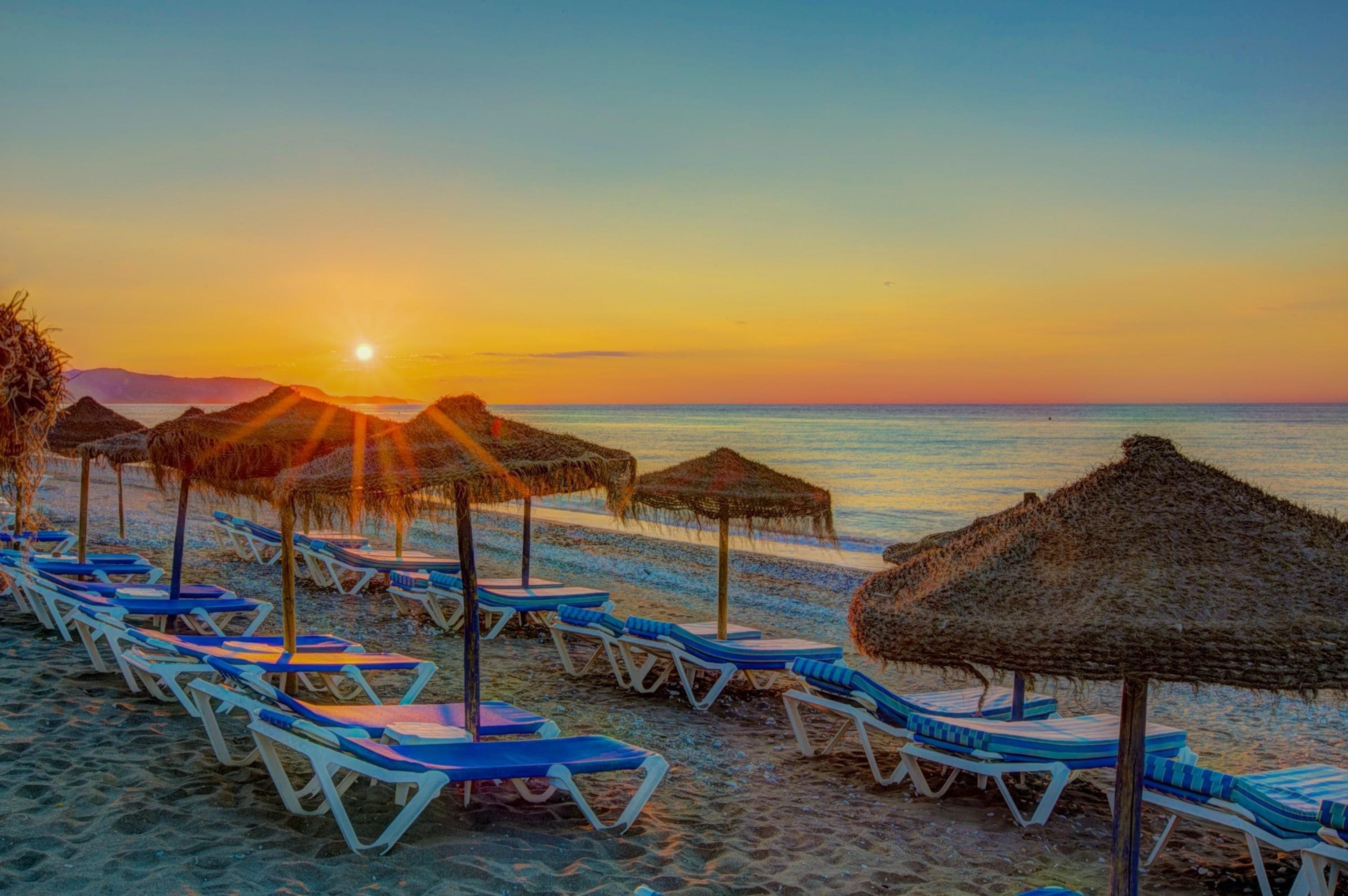 Ein Strand mit Liegestühlen und Reetdachschirmen, im Hintergrund ein Sonnenuntergang und ruhige See.