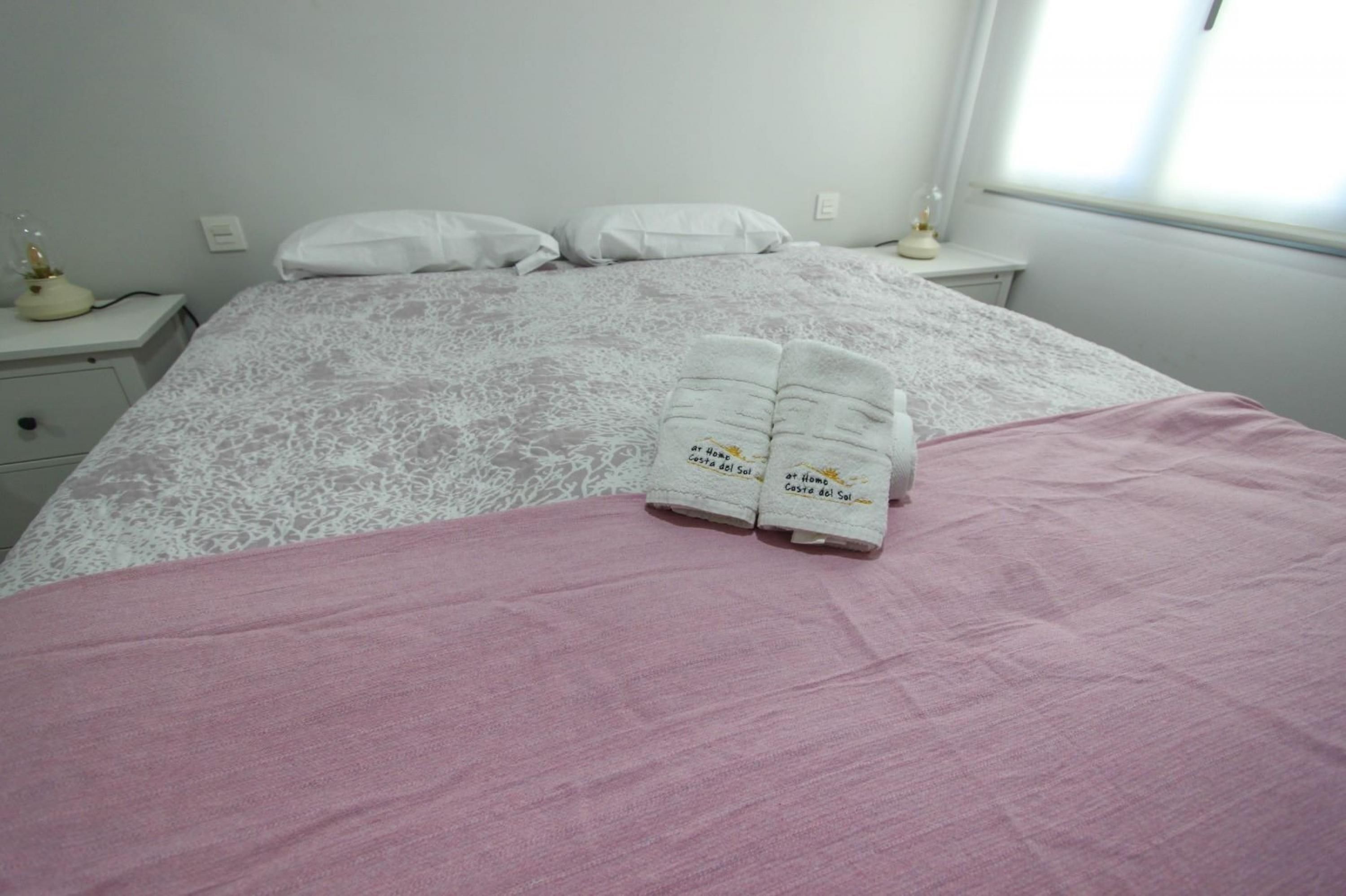 Een net opgemaakt bed met een roze deken en twee handdoeken met tekst erop.