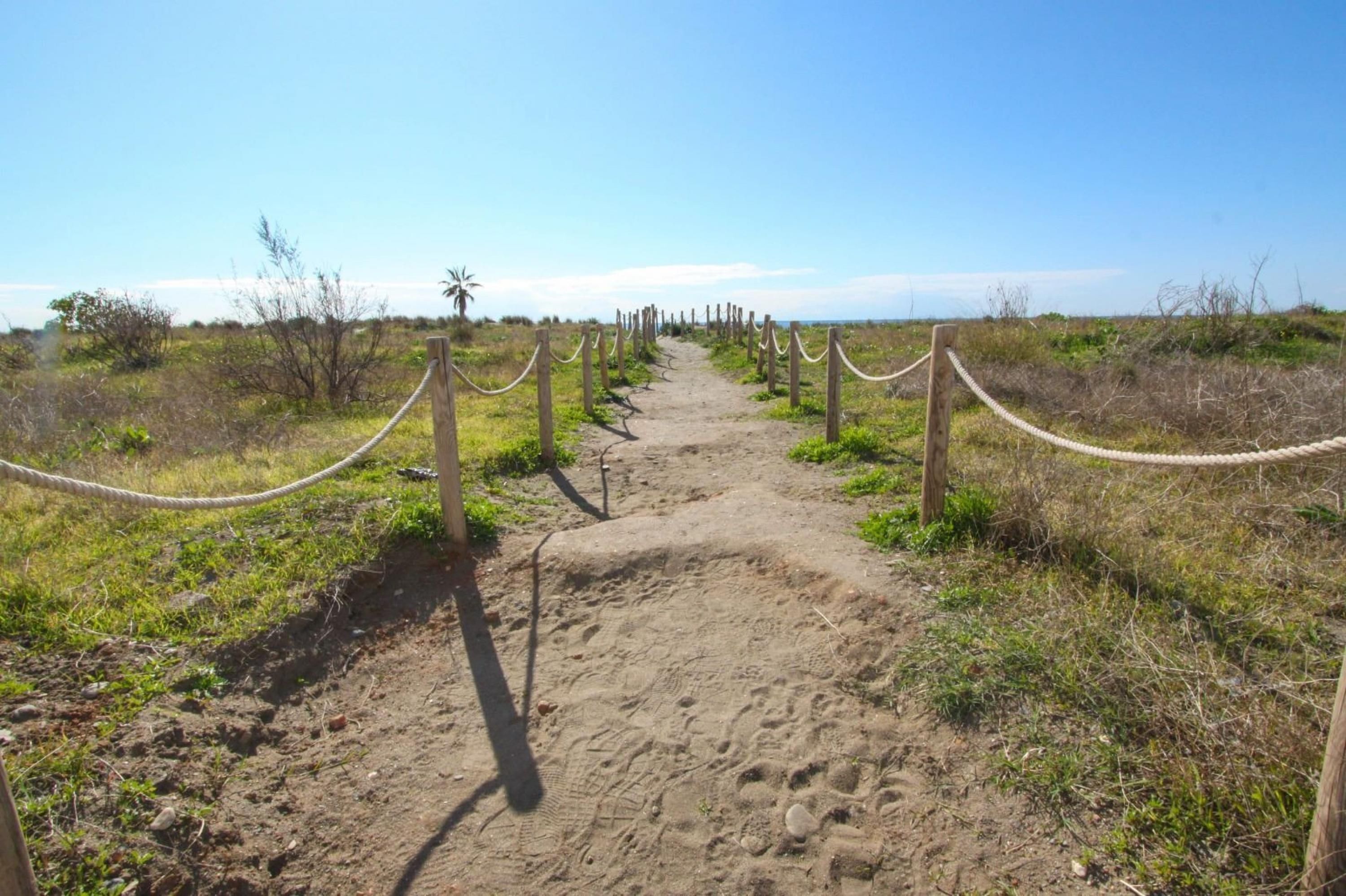 A sandy path with wooden posts and ropes, leading towards a natural landscape.