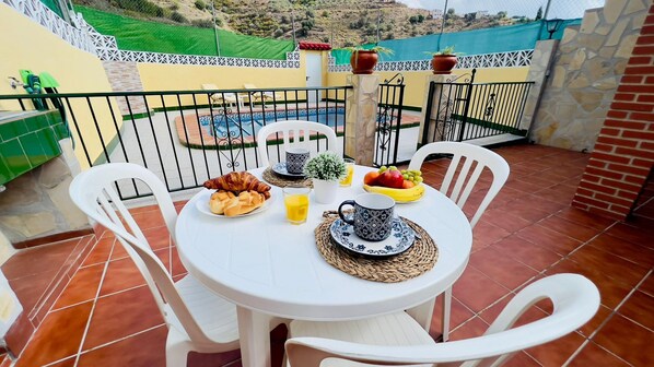 Uma mesa redonda com cadeiras brancas, um ambiente de café da manhã com croissants, frutas e sucos, e vista para uma piscina e vegetação ao fundo.