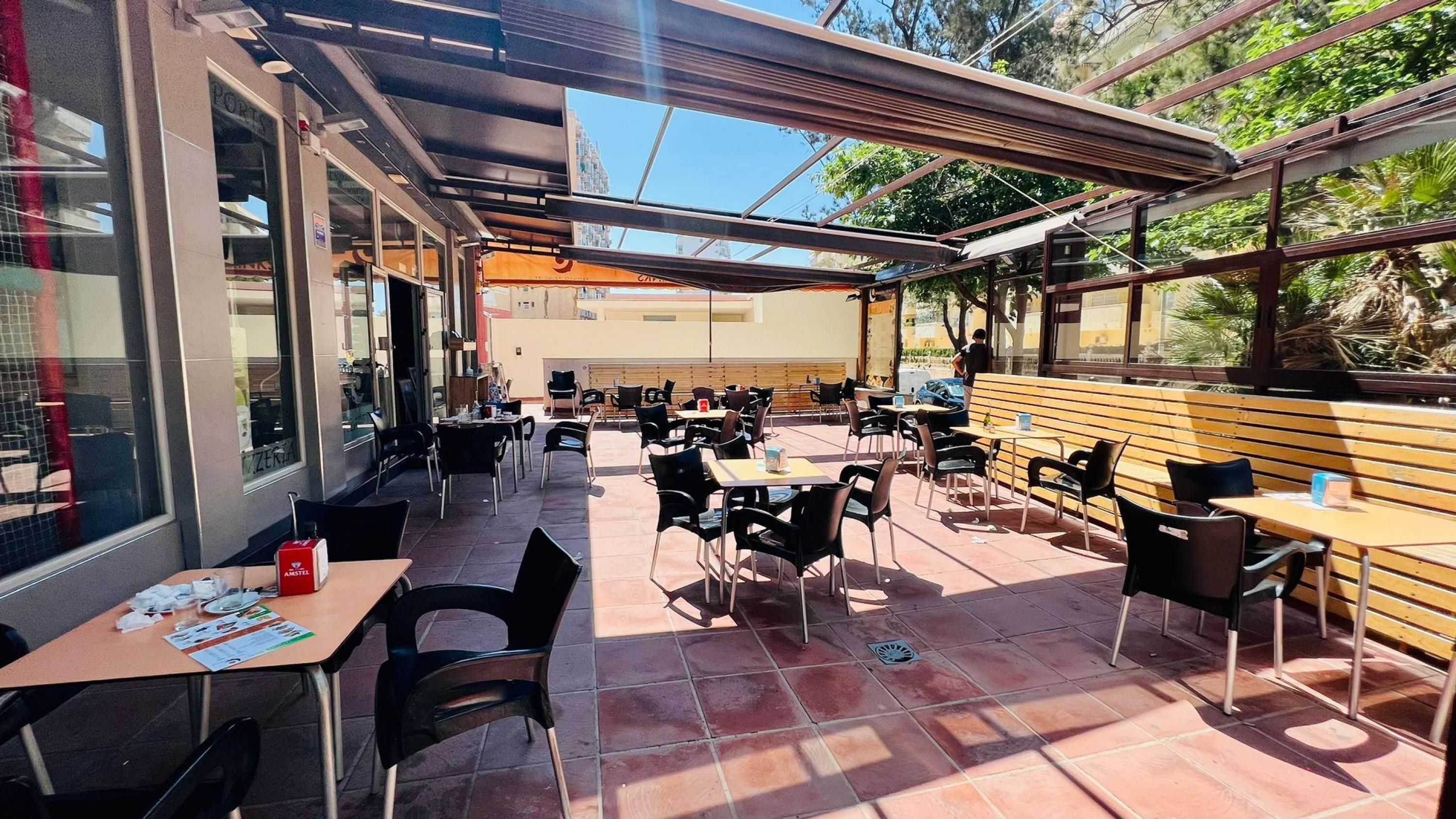 An outdoor cafe with tables and chairs, a wooden bench, and a glass wall.