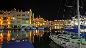 Un port de plaisance de nuit, avec des bateaux amarrés, des bâtiments illuminés et une surface d’eau calme qui reflète les lumières.
