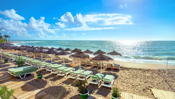 Une plage bordée de rangées de chaises longues et d’ombrelles en chaume, une terrasse en bois et une vue sur l’océan.
