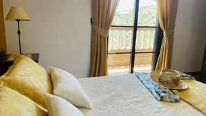 Een bed met een witte quilt en twee kussens, een lamp, een geweven mand en uitzicht op een balkon met gordijnen.