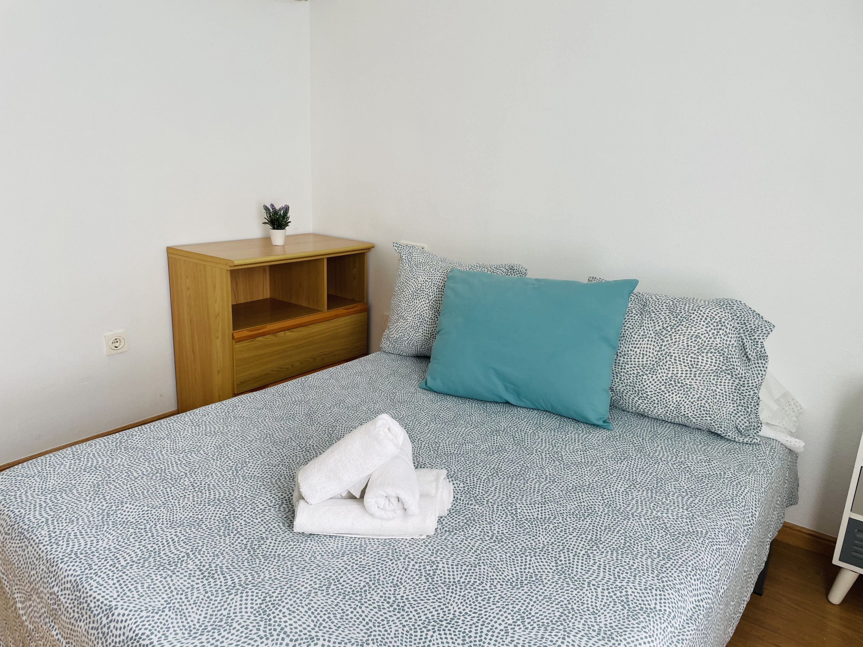 A neatly made bed with patterned bedding, a teal pillow, and rolled towels on the bed.