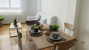 Un comedor con mesa de madera, sillas y sofá blanco. Hay plantas en macetas, un cuenco de fruta y una mesa baja con un adorno.