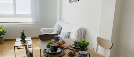 A dining area with a wooden table, chairs, and a white sofa. There are potted plants, a bowl of fruit, and a coffee table with a decorative item.