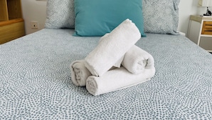 Una cama bien tendida con ropa de cama estampada, una almohada verde azulada y toallas enrolladas.