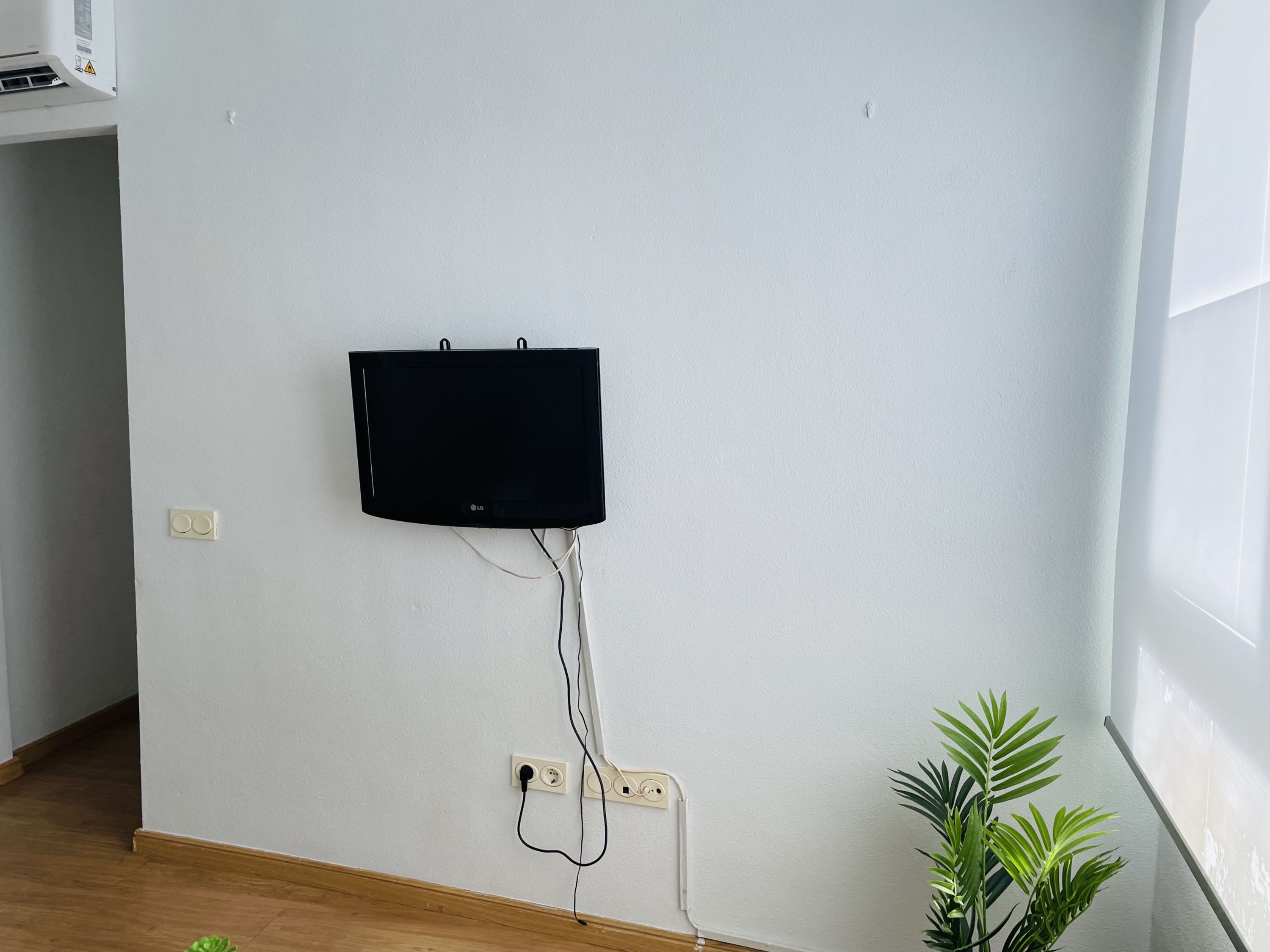 A wall-mounted flat-screen TV with visible power cables, a wall socket, and a potted plant in a room with white walls and wooden flooring.