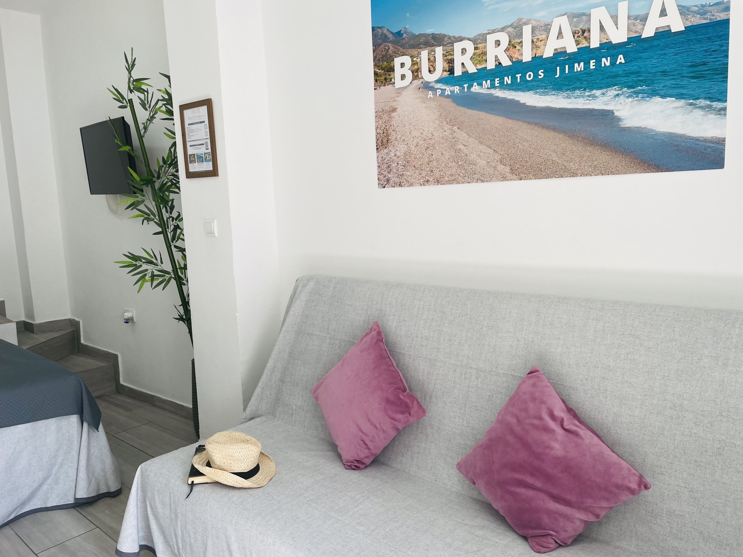 A modern living room with a grey sofa, two pink cushions, and a straw hat on the seat. A poster of a beach scene is on the wall.