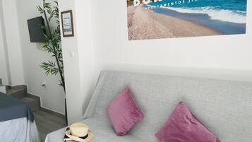 A modern living room with a grey sofa, two pink cushions, and a straw hat on the seat. A poster of a beach scene is on the wall.