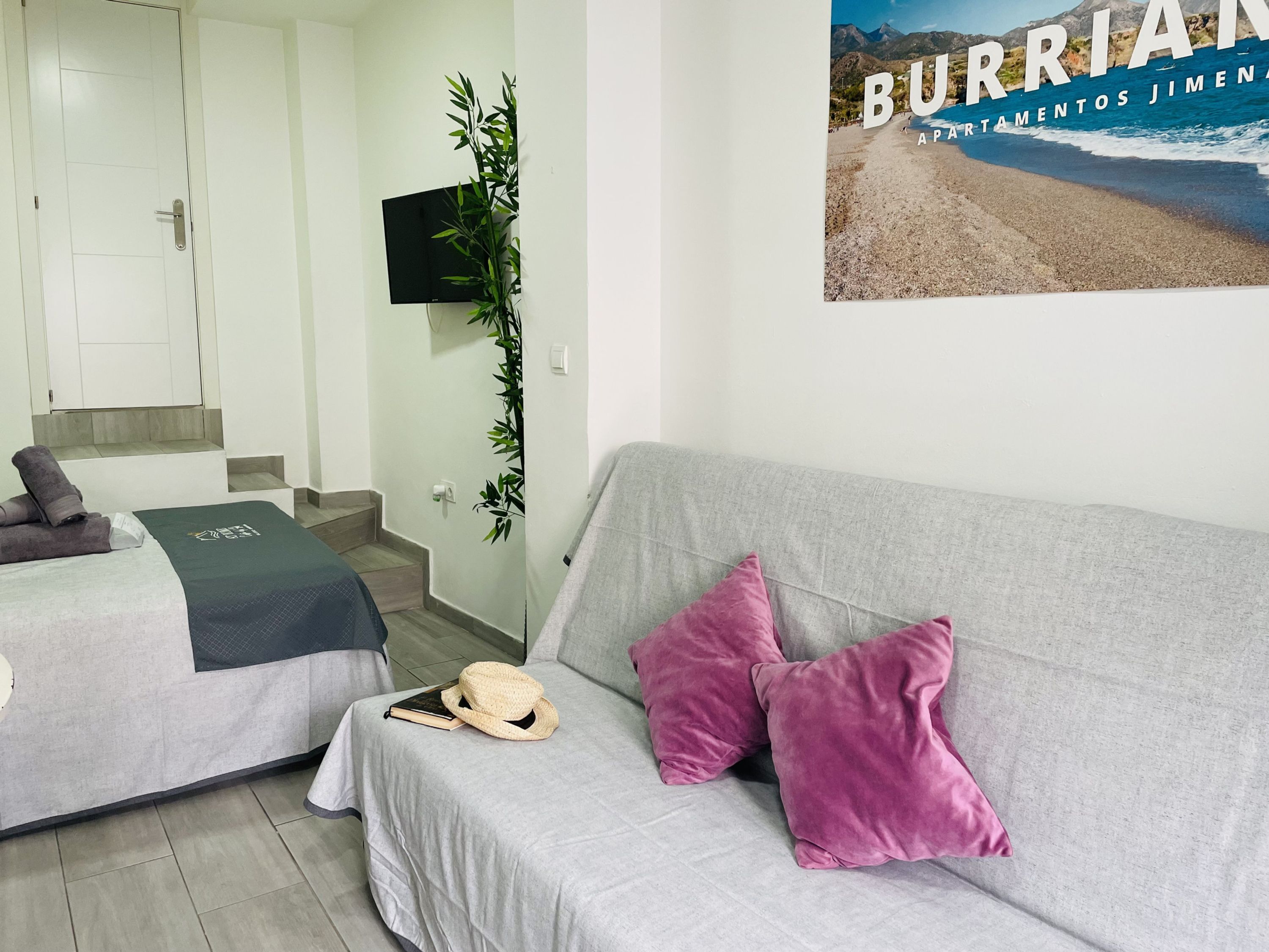 A hotel room with a grey sofa, two beds, a TV, a poster of a beach, and a staircase.