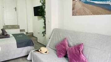 A hotel room with a grey sofa, two beds, a TV, a poster of a beach, and a staircase.