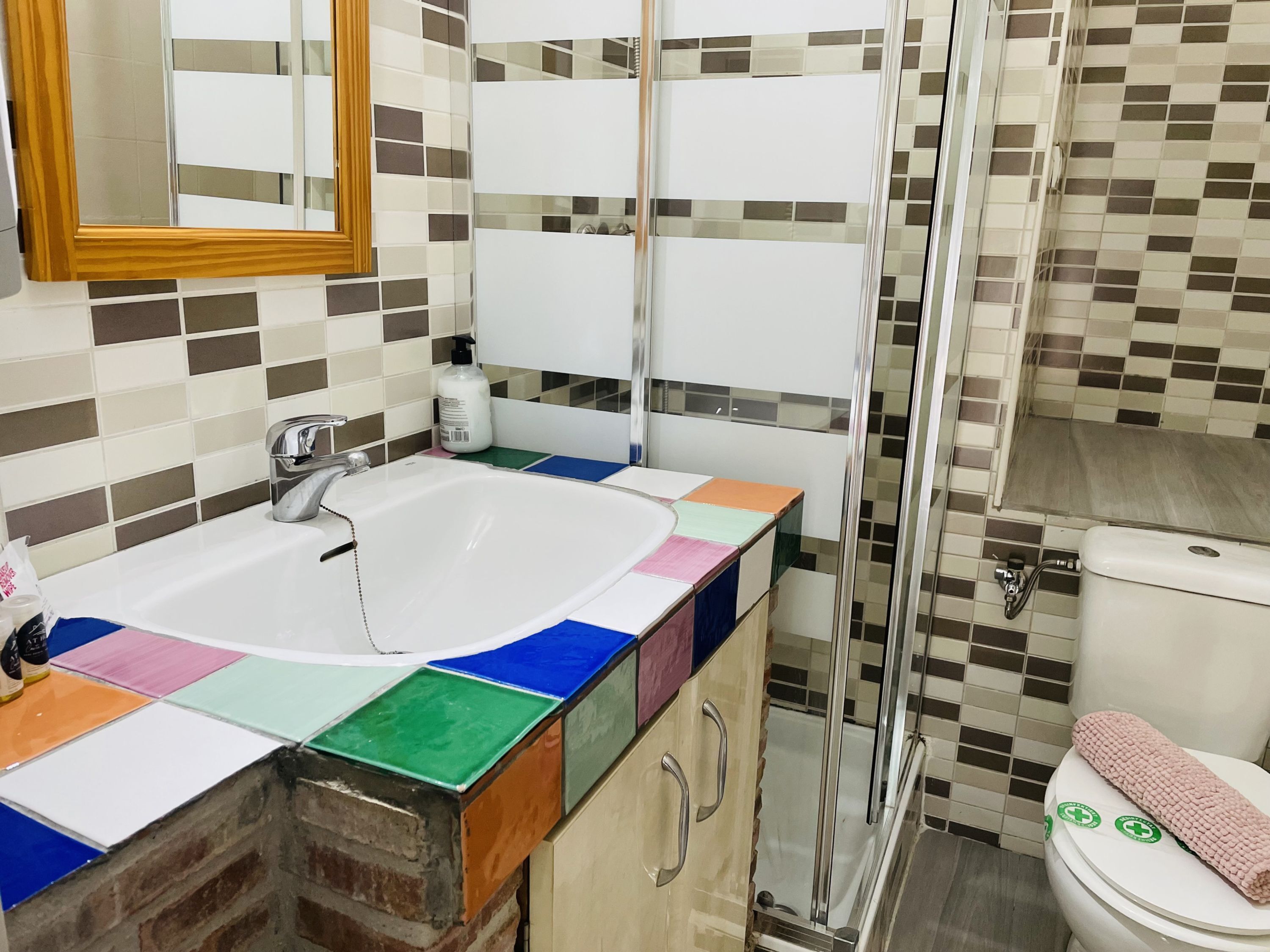 Une salle de bain avec un lavabo carrelé, un miroir, des toilettes et un espace douche.