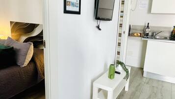 Un espacio habitable pequeño con cama, una mesita blanca, televisor montado en la pared, zona de cocina con lavamanos y encimera, y ducha.