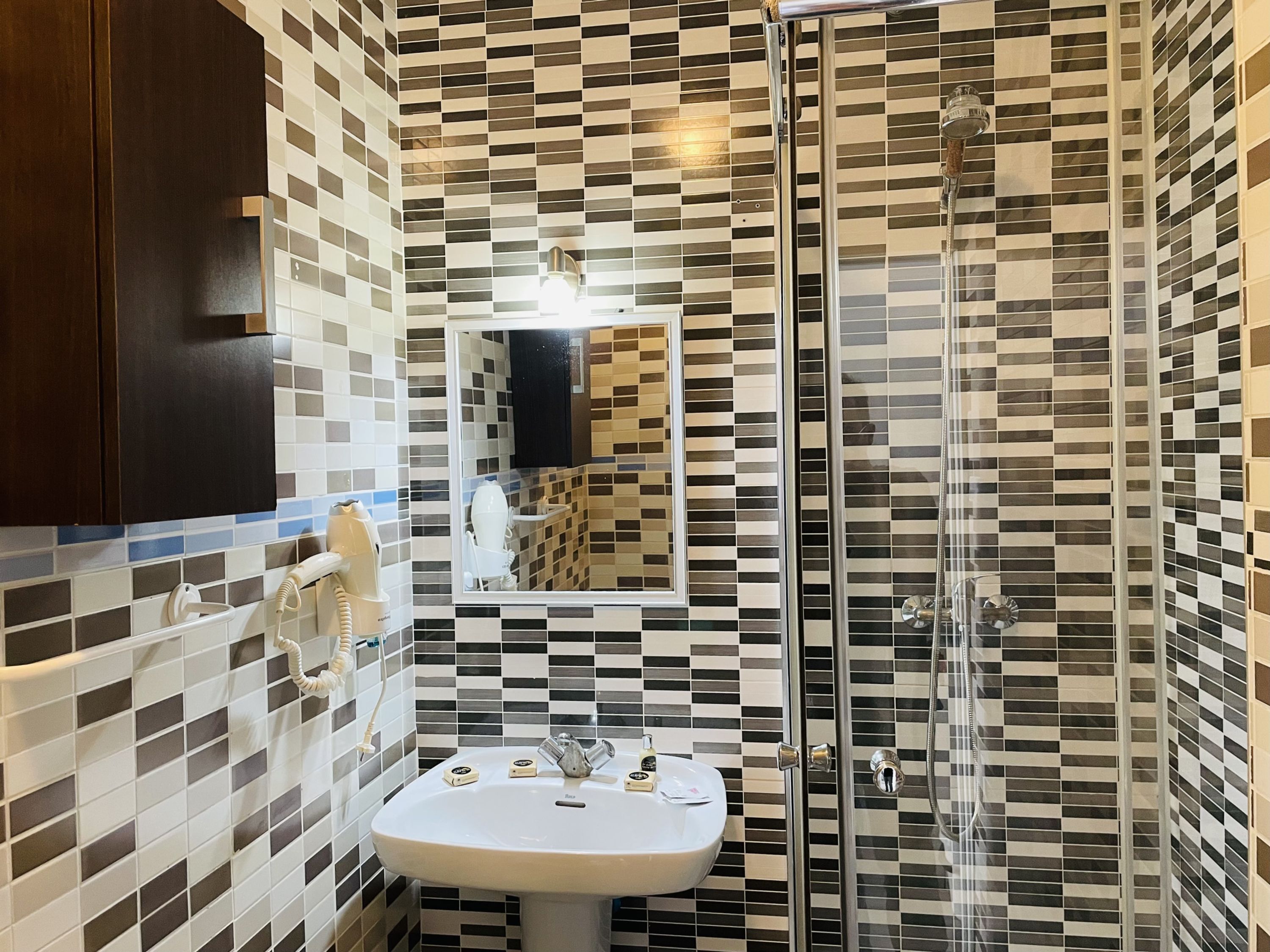 A bathroom with a tiled wall, a sink, a mirror, a shower, and a telephone.