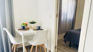 Quarto pequeno e moderno com uma mesa de jantar, duas cadeiras brancas, uma cama e uma porta que dá para outro espaço.