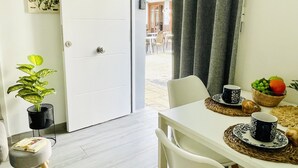 Um quarto com uma porta branca, um vaso de planta, uma mesa de jantar com uma cesta de frutas e uma chávena, e uma cadeira.
