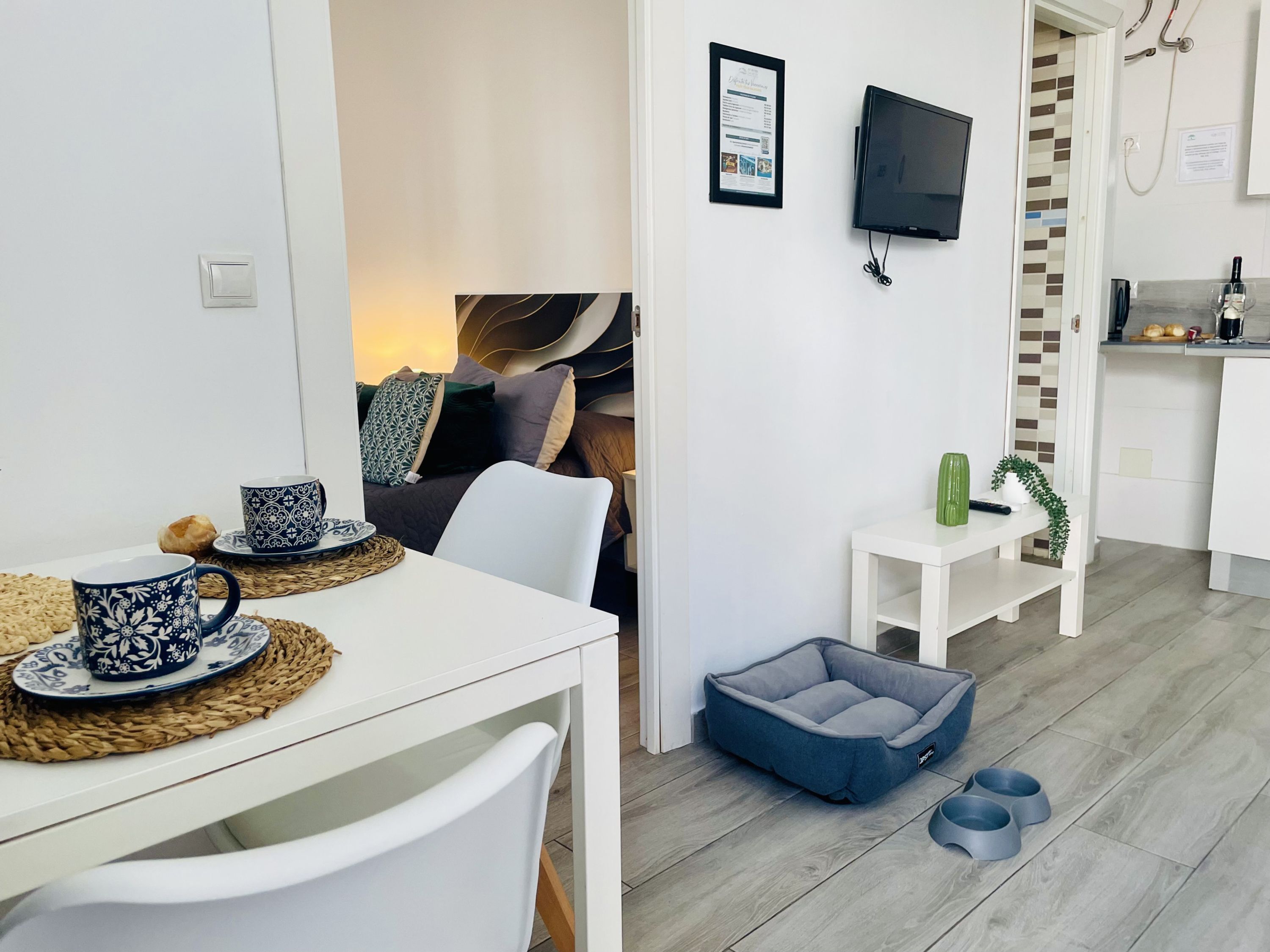 Una sala moderna con un sofá blanco, una mesa de comedor con vajilla de estampado azul, una cama gris para mascotas, un televisor montado en la pared y una pequeña cocina.