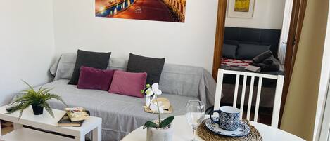 A cozy living room with a sofa, a coffee table, and a dining table set with a book, a plant, a glass, and a fruit platter.