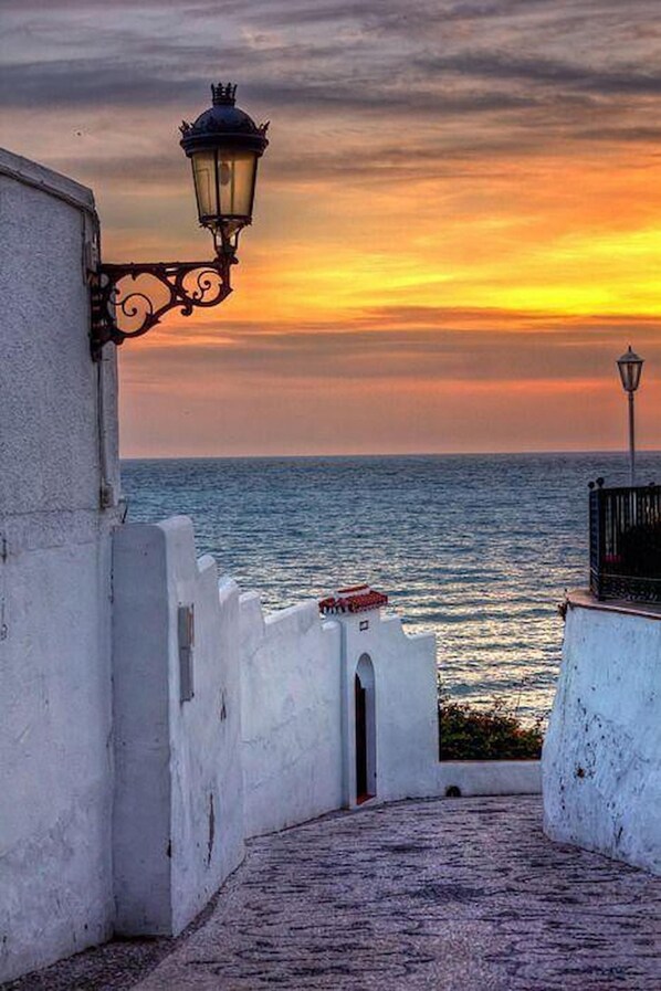 A coastal pathway with a white building, a street lamp, and a view of the sea at sunset. - Apartamento Malaga Jimena by At Home Costa del Sol (Nerja)