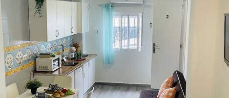 A compact kitchen with white cabinets, a microwave, and a small dining area with a table and chairs. There is a sofa with orange pillows in the adjacent living area.