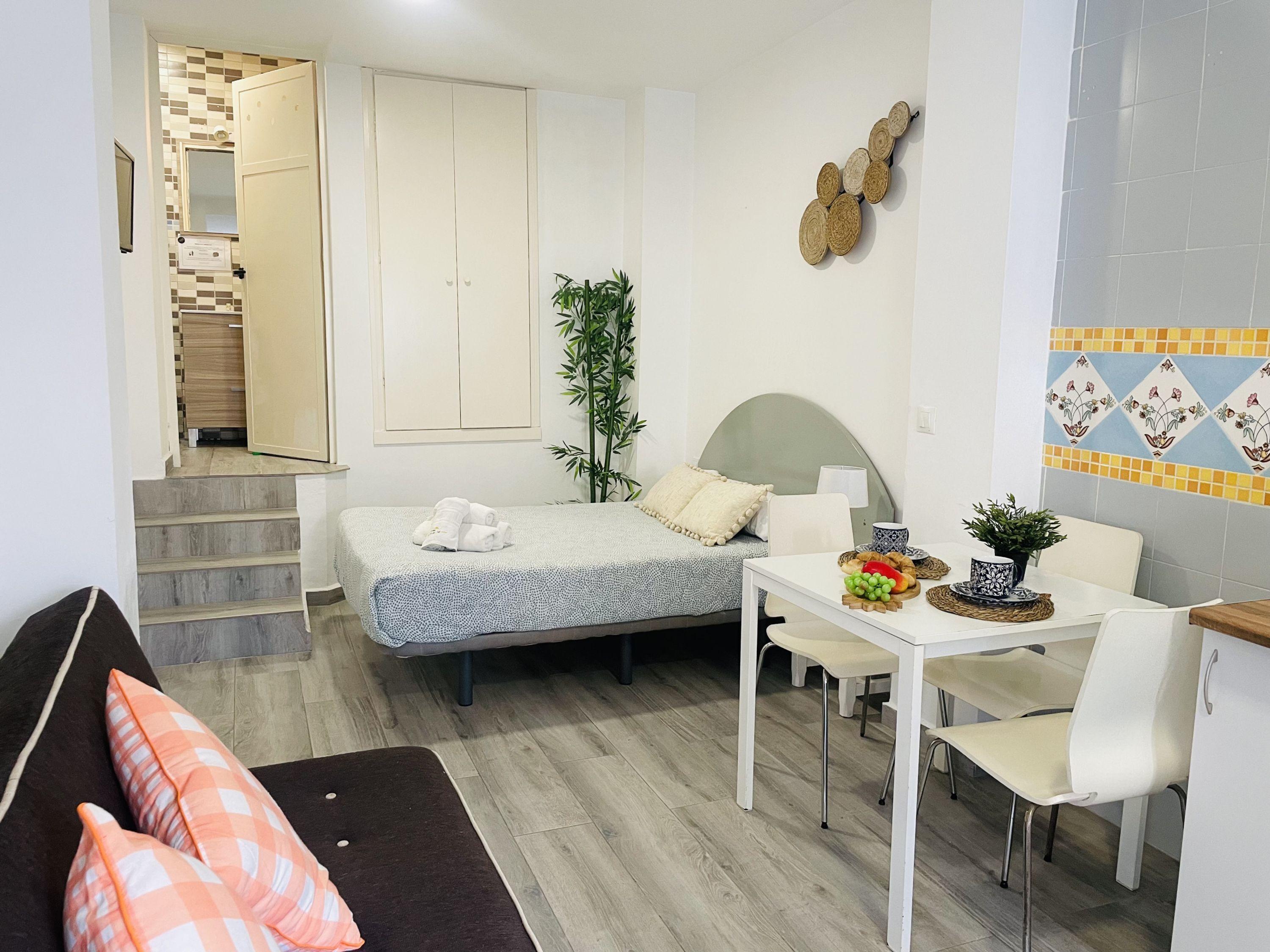 A compact hotel room with a bed, a dining table with chairs, a small staircase, and a plant.