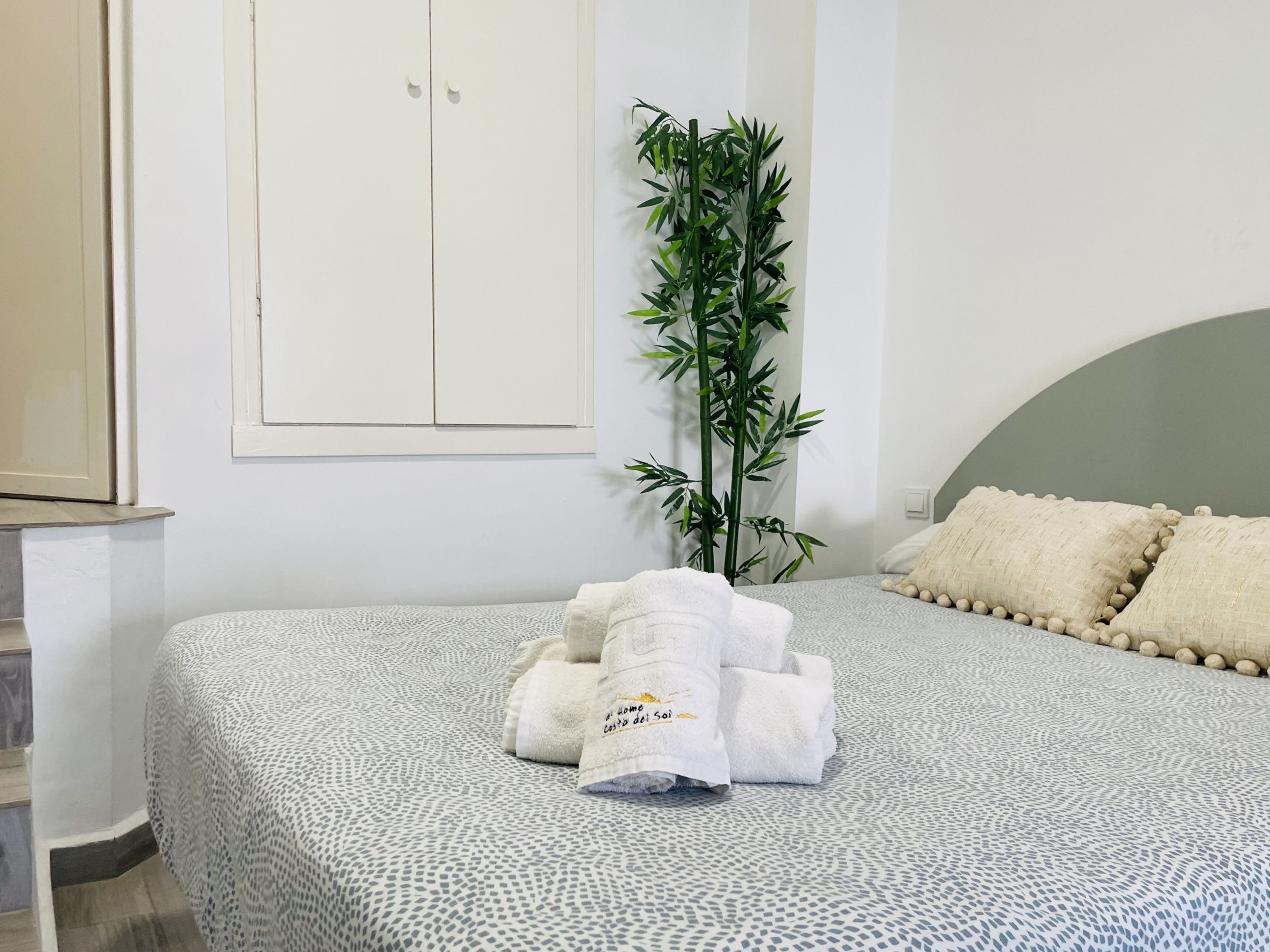 A neatly made bed with a patterned bedspread, a set of towels, a tall green plant, and a white wardrobe.