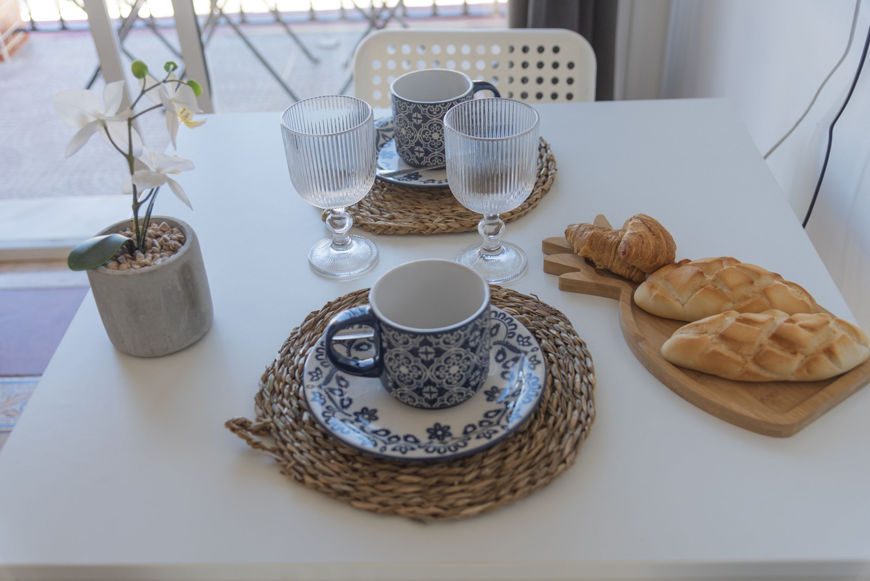 Une table dressée avec deux tasses, deux verres à vin, une assiette de croissants et une orchidée en pot.