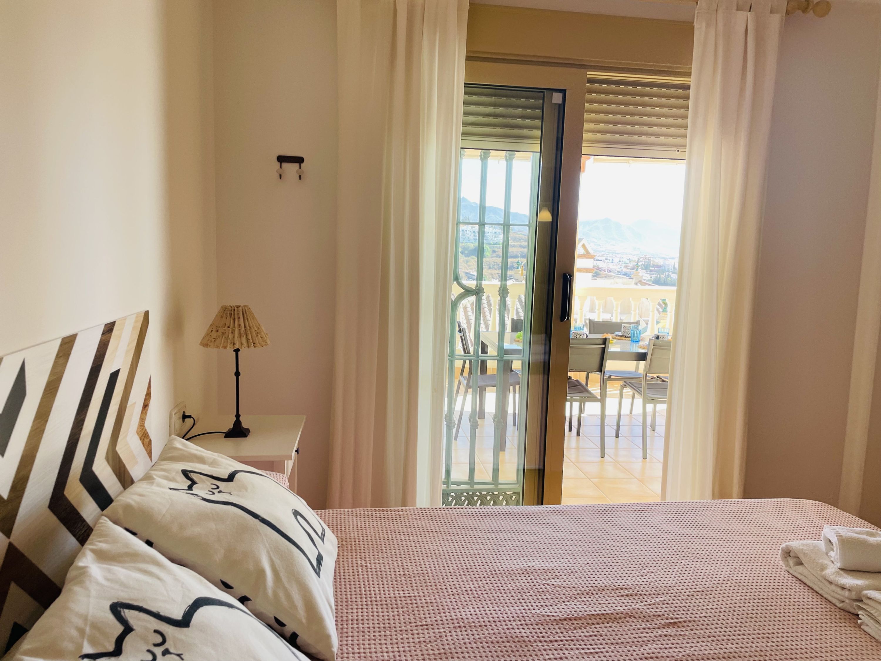 A hotel room with a bed, a nightstand, a lamp, and a view of a balcony with outdoor seating.