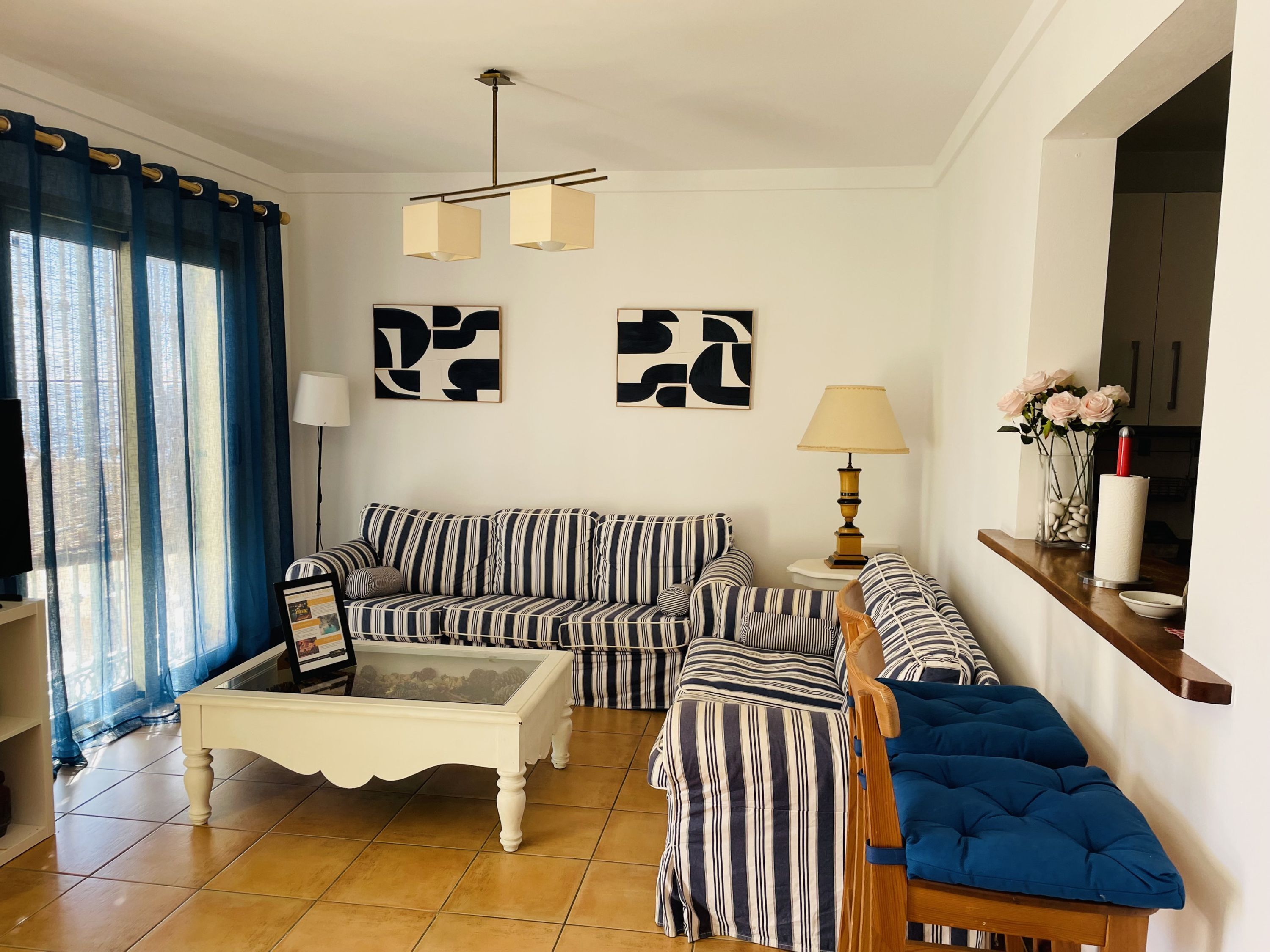 A living room with a striped sofa, a coffee table, and a blue ottoman. There are abstract wall art pieces and a lamp on a side table.
