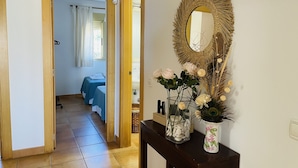 Une chambre d’hôtel avec un lit, une salle de bain et un couloir menant à une autre pièce. Un miroir avec un cadre décoratif est accroché au mur, et un vase rempli de fleurs est posé sur une table.