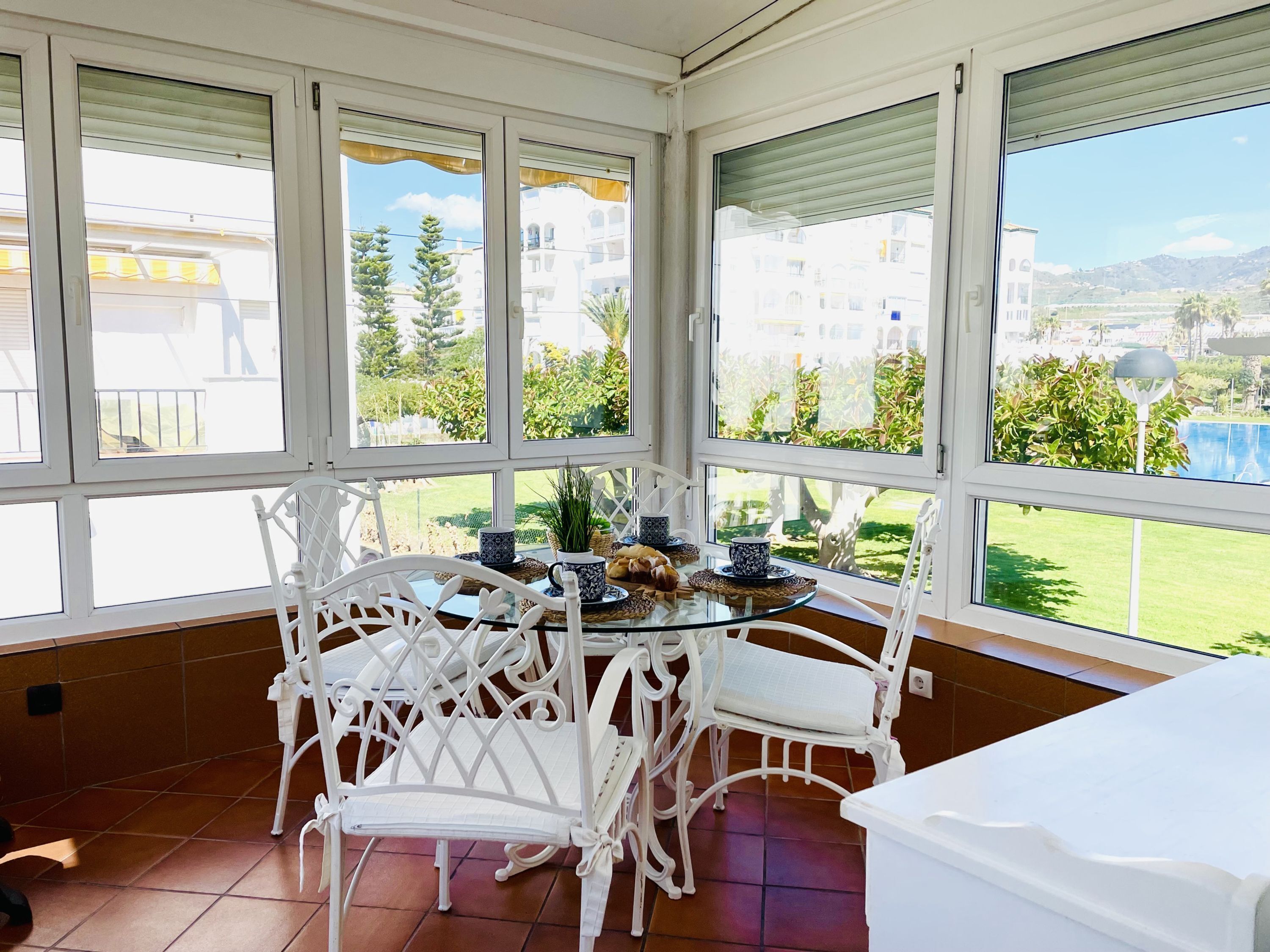 A sunroom with a glass table and white chairs, overlooking a green landscape and buildings.