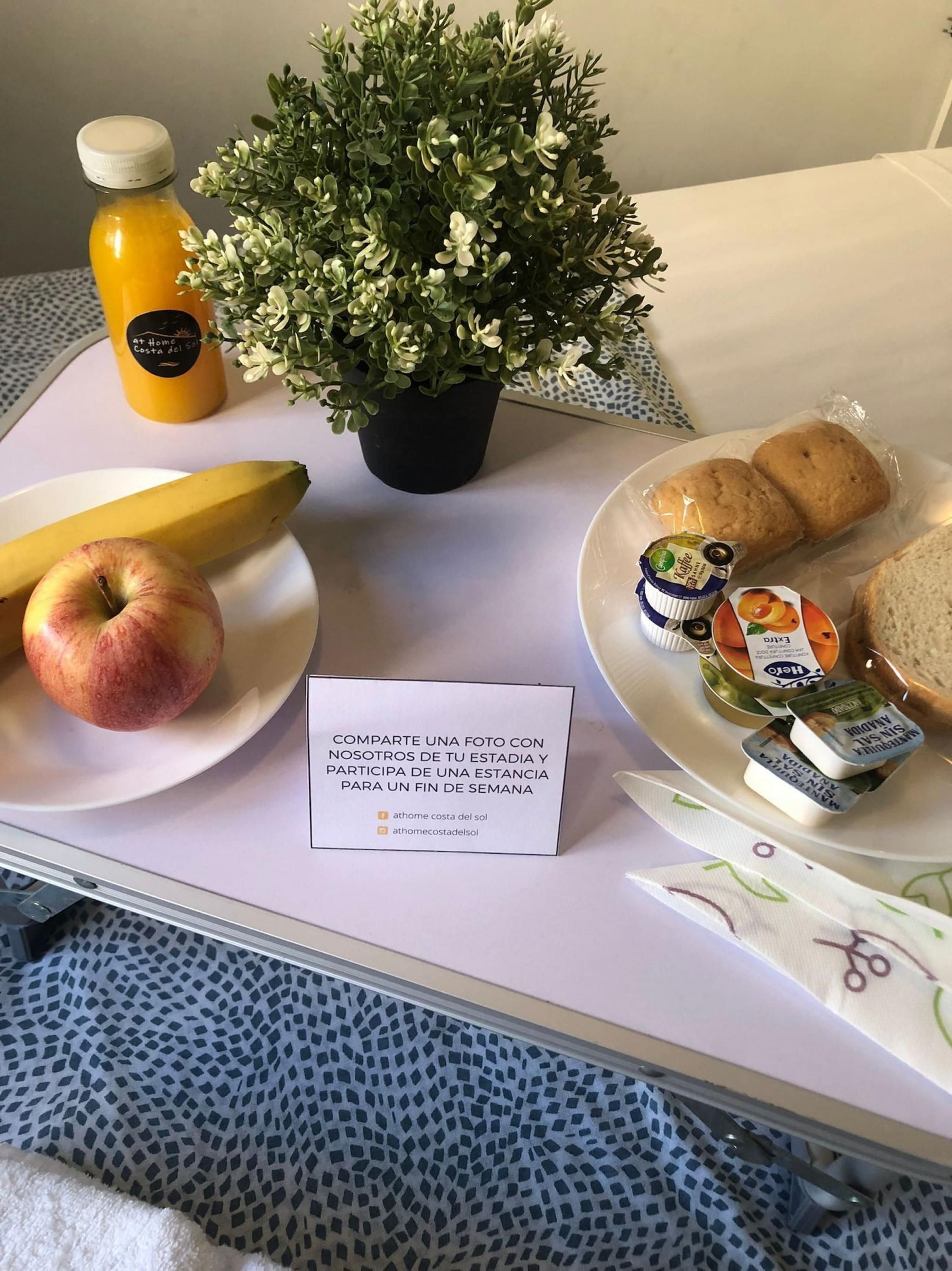 A tray with a bottle of orange juice, a banana, an apple, a potted plant, a plate with biscuits and a jar of jam, and a note inviting photo sharing.