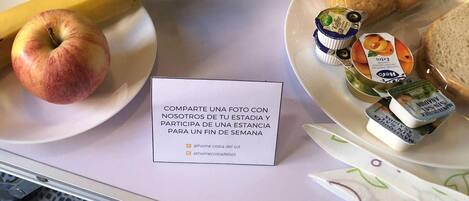 A tray with a bottle of orange juice, a banana, an apple, a potted plant, a plate with biscuits and a jar of jam, and a note inviting photo sharing.