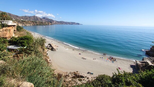A coastal beach with clear blue waters, rocky cliffs, and a few buildings on the left. - Apartamento Huelva Jimena by At Home Costa del Sol (Nerja)