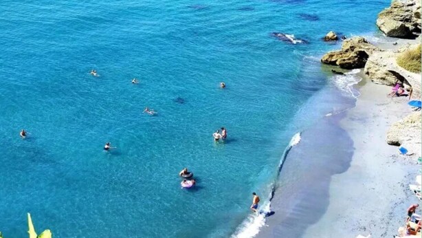 A beach with clear blue water, people swimming and sunbathing, and rocky terrain.