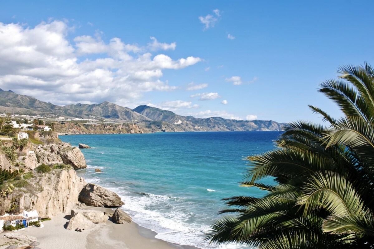 A coastal landscape with a beach, rocky cliffs, and a mountain range in the background.