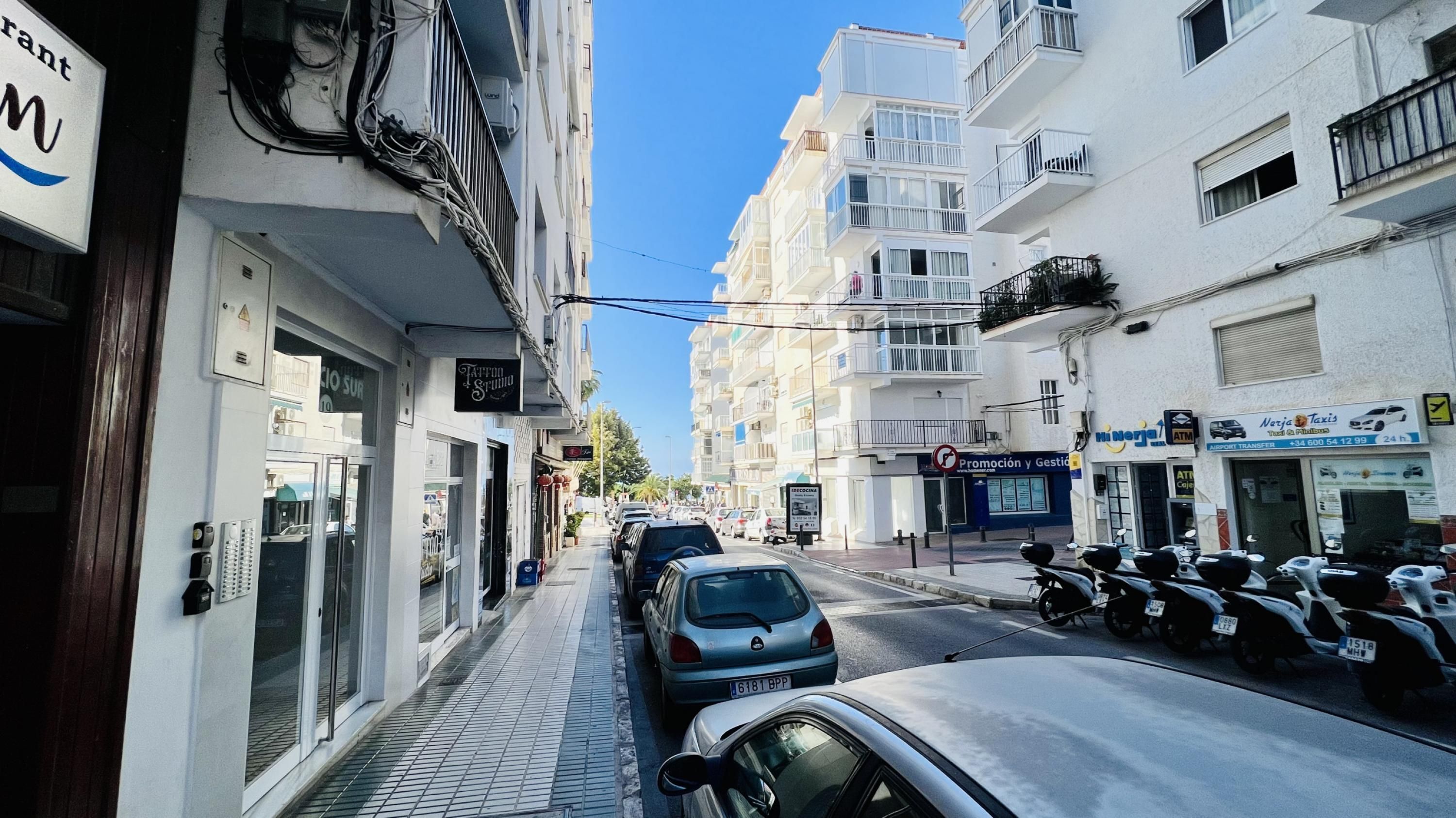 A narrow street lined with multi-story buildings, some with balconies, and parked cars and scooters.