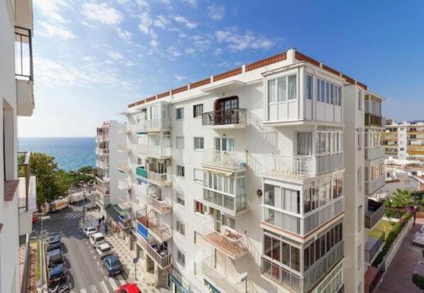 A multi-story residential building with balconies, a view of the sea, and parked cars on the street.