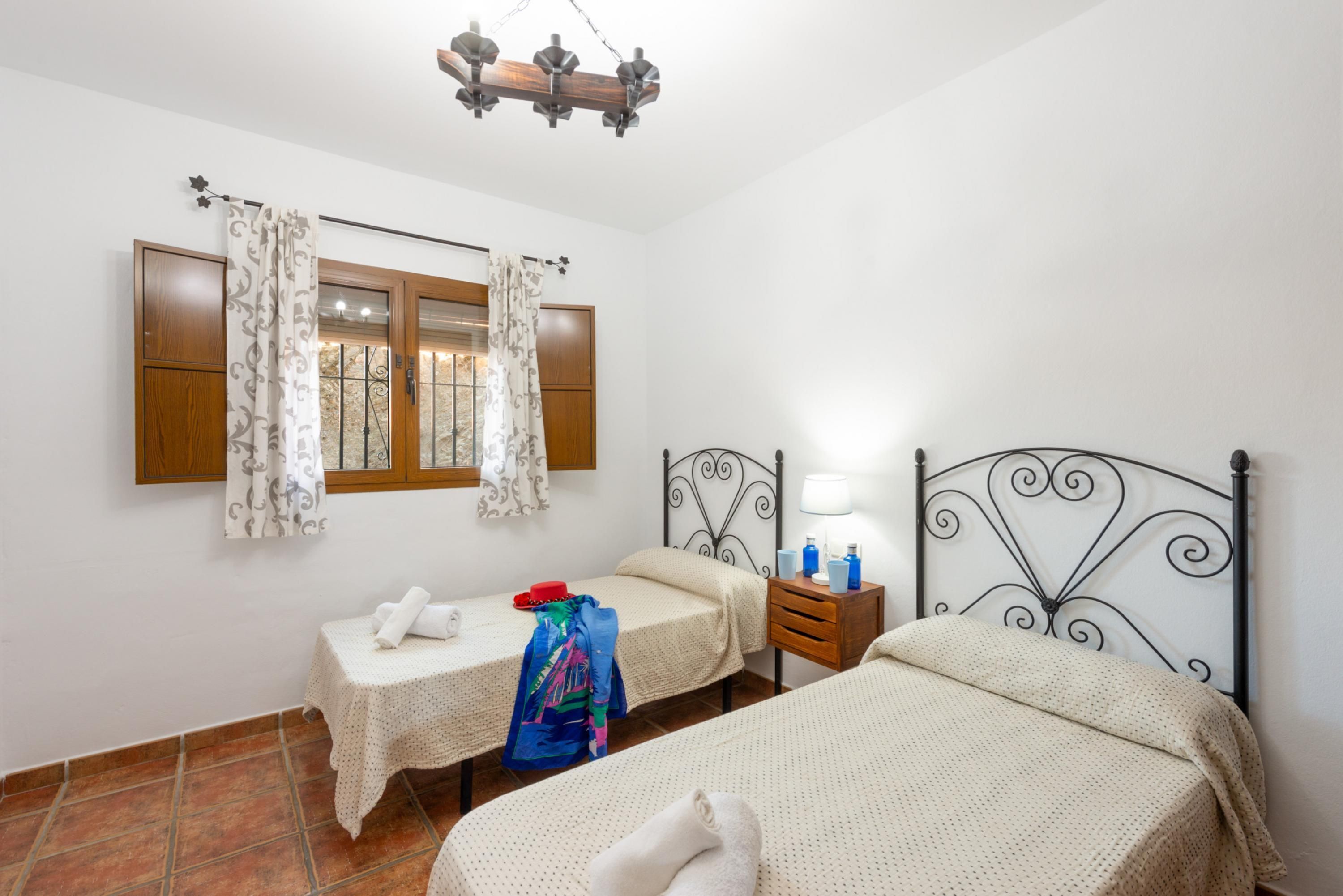 A bedroom with two beds, a window with curtains, a bedside table with a lamp, and a wooden headboard.