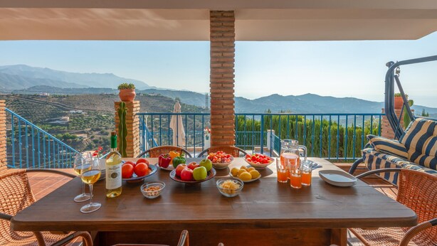 Een houten tafel gedekt met fruit, groenten en drankjes op een balkon met uitzicht op een berg.