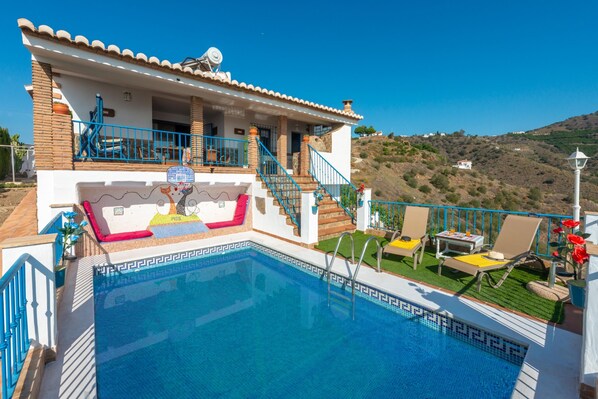 A poolside area with a clear view of the pool, a covered seating area with chairs and a table, and a staircase leading to the upper floor.