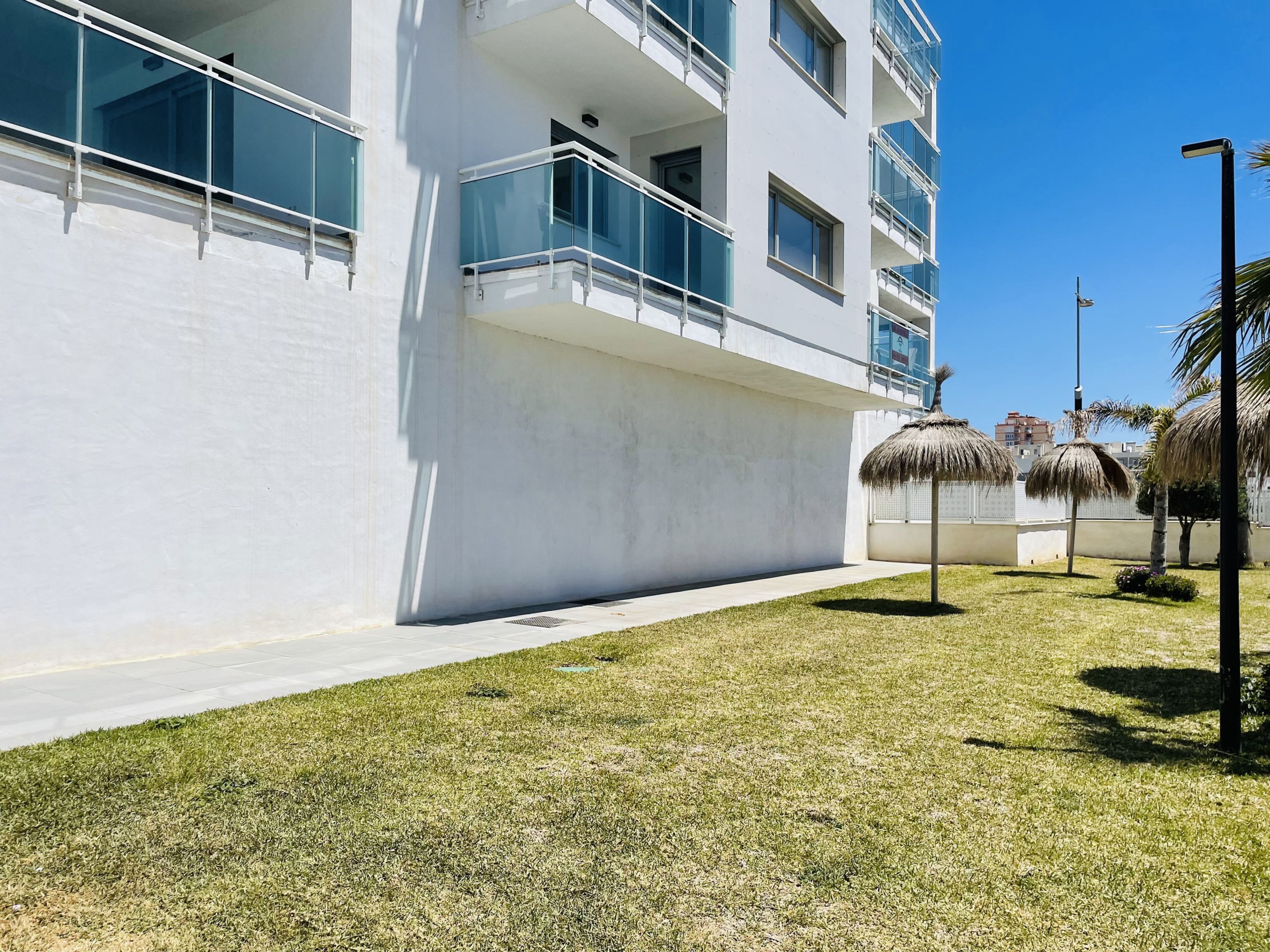 Un bâtiment moderne doté de balcons, d’une allée pavée et d’un espace verdoyant agrémenté de parasols en chaume.